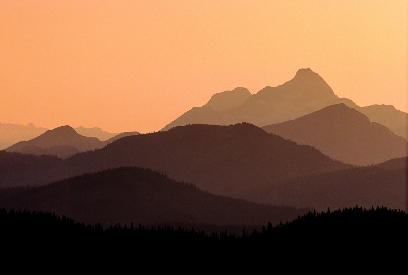 Layered mountain ridges in silhouette at sunset with a warm orange sky.